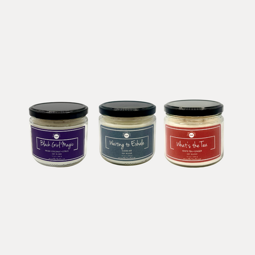 Three 12 oz glass jar candles with cotton wicks and soy blend wax: one with a purple label reading 'Black Girl Magic,' another with an orange label reading 'What's the Tea,' and a third with a bluish-gray label reading 'Waiting to Exhale.' Perfect for self-care or as a thoughtful gift for a Black woman.