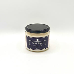 12 oz glass jar candle with cotton wicks and soy blend wax, featuring a non-toxic fragrance. Designed to evoke feelings of pride, self-worth, and nostalgia. Midnight blue label reads 'Harlem Nights'. Fragrance combines violet, amber, and musk for a powdery night-time scent.